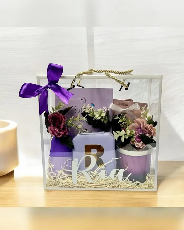 This personalized gift for 'Ria' is presented in a stunning transparent acrylic bag with a purple ribbon. It features a custom initial on a travel case and other curated items in a matching lavender theme.