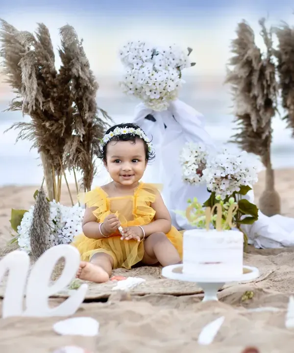 A beautiful first birthday setup on the beach, with a baby girl in a yellow dress sitting beside her cake, with the ocean in the background.
