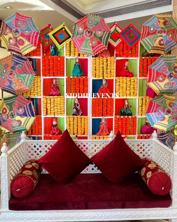 We created this joyful Haldi backdrop using colorful Gujarati umbrellas, marigold garlands, and traditional Rajasthani dolls, all set against a white diwan with rich red cushions.