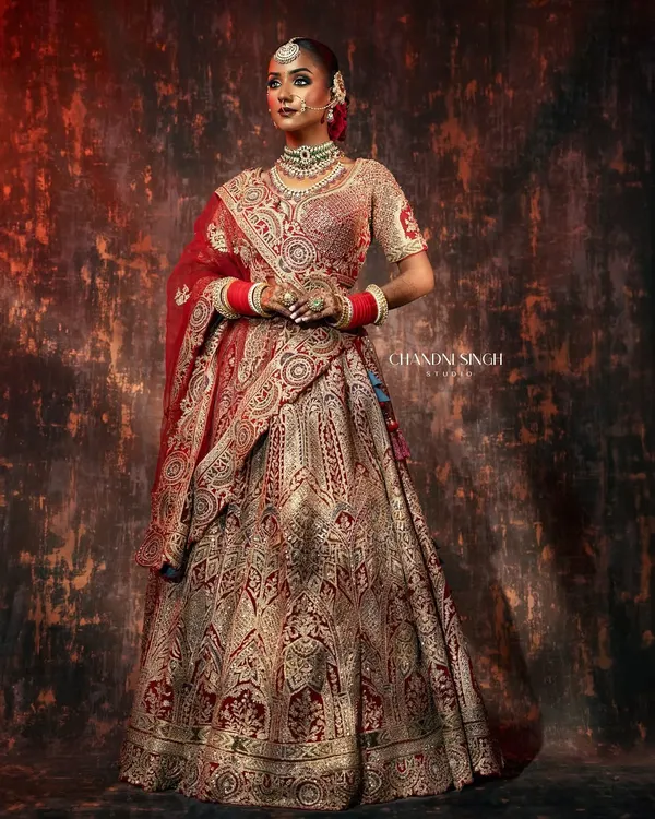 A full-length bridal portrait showcasing a magnificent red and gold lehenga. The lighting is sculpted to highlight the rich embroidery and create a regal, statuesque presence.