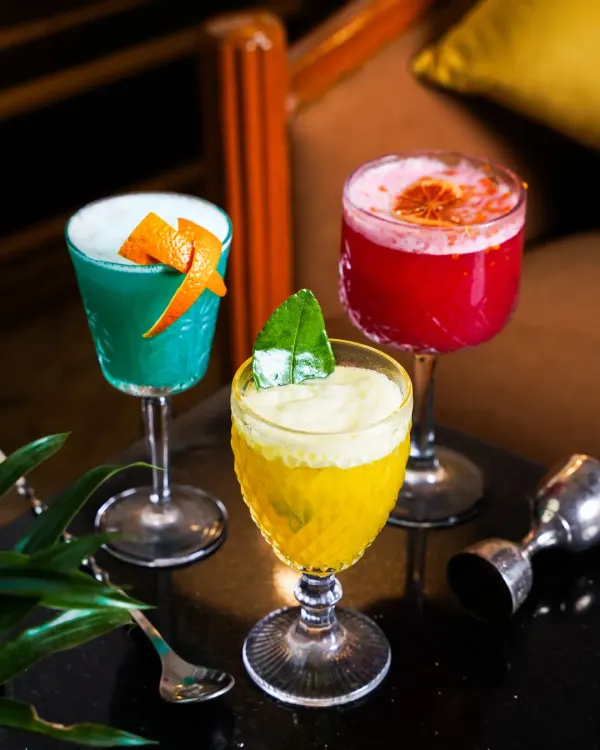 A trio of colorful cocktails, including a blue curaçao drink, a vibrant red concoction, and a frothy yellow sour, styled for a moody bar setting. This shot is perfect for showcasing a diverse cocktail menu.