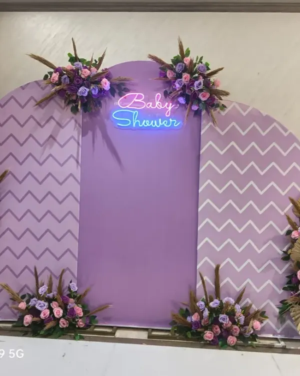 A modern boho baby shower backdrop in lavender and white. This design features chevron-patterned panels, a 'Baby Shower' neon sign, and beautiful arrangements of purple flowers and dried golden leaves.