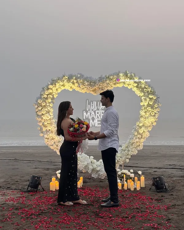I can help you create the perfect moment to ask the big question. This beach proposal setup features a large, heart-shaped arch made of white flowers, illuminated to stand out against the evening sky, with a "Will You Marry Me?" neon sign and a pathway of rose petals.