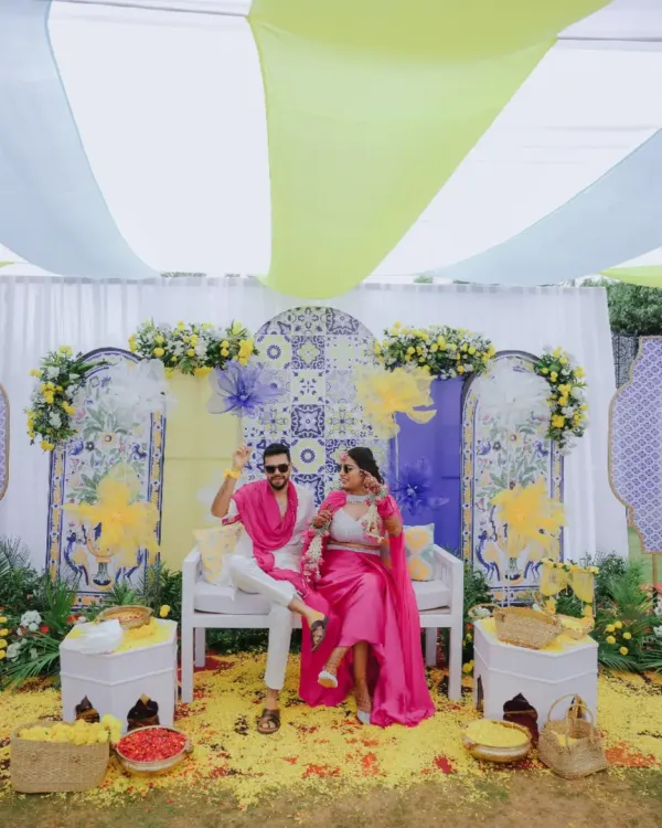 A vibrant Haldi setup featuring a backdrop of blue and yellow tiles, framed by bright pink outfits and a shower of petals. I love creating a playful atmosphere for this fun-filled ceremony.