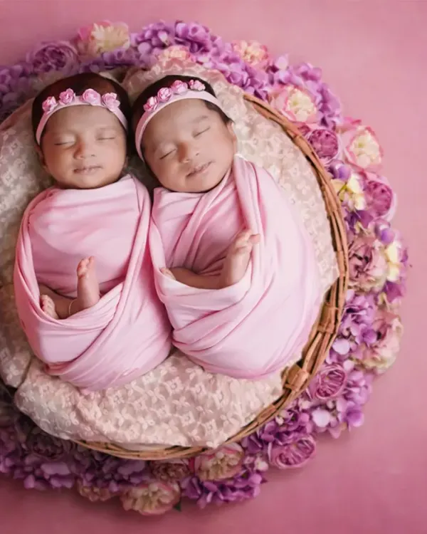 The perfect pair. Two newborn twin girls swaddled in pink, sleeping side-by-side in a basket surrounded by purple flowers. This is a classic and heartwarming twin portrait.