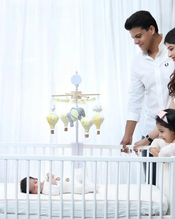 A heartwarming lifestyle scene where parents and an older sister gather around the crib to watch over the family's newest member. The all-white outfits create a clean, serene atmosphere.