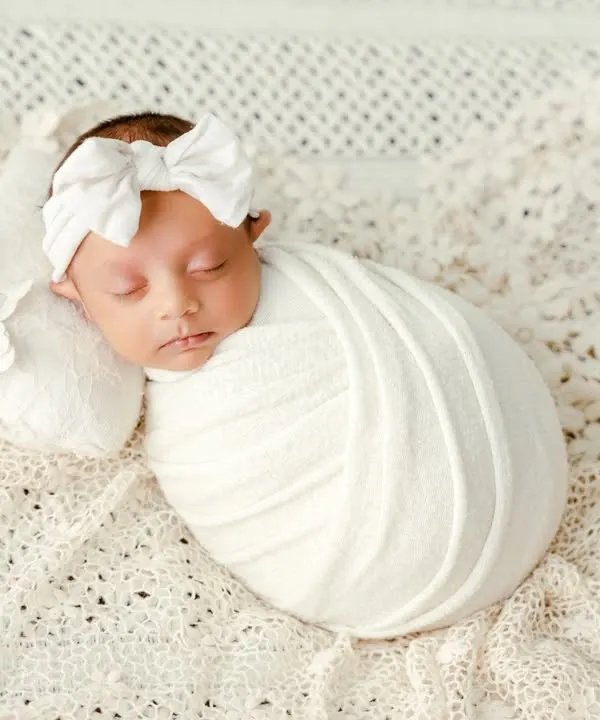 A classic newborn portrait focusing on purity and simplicity. The baby is swaddled snugly in white fabric on a soft, textured blanket, creating a timeless and angelic look.