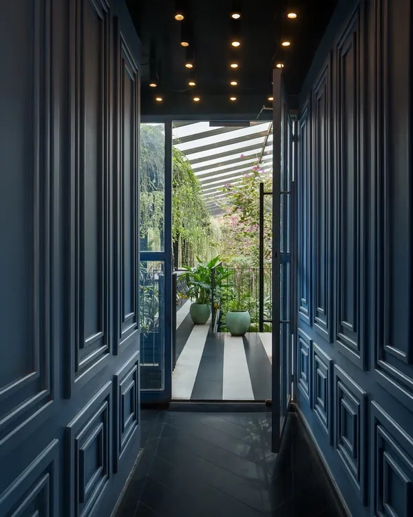 A doorway framed by deep blue paneled walls offers a glimpse from a moody corridor into the bright, plant-filled garden court. This transition highlights the deliberate contrast I create between different zones to evoke distinct emotional responses.