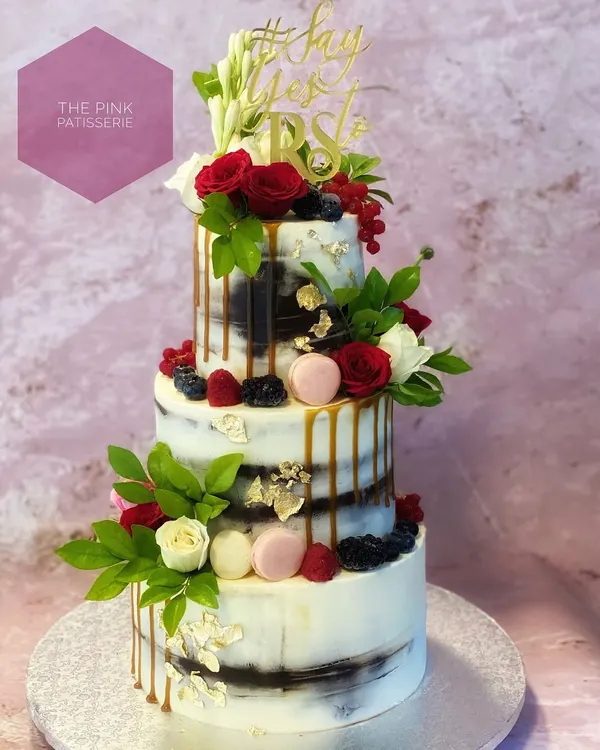 A three-tiered semi-naked cake for a proposal, topped with a "She Said Yes" acrylic sign. This rustic yet elegant design is adorned with fresh red and white roses, berries, and a caramel drip.