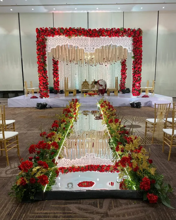 A grand entrance for a classic red and white wedding. The mirrored aisle is lined with red and yellow flowers, leading to a magnificent mandap that promises a beautiful ceremony.