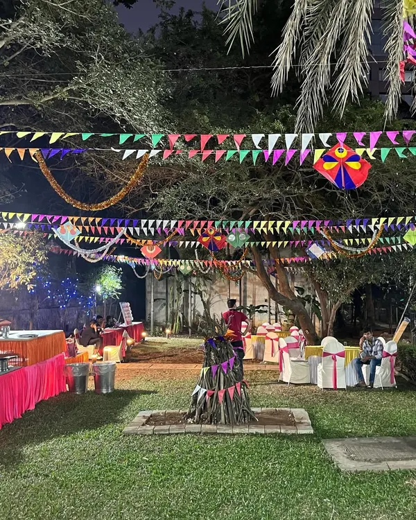 An outdoor Lohri celebration at night. The garden is decorated with colorful flags, kites, and traditional hangings, with a bonfire set up in the center.