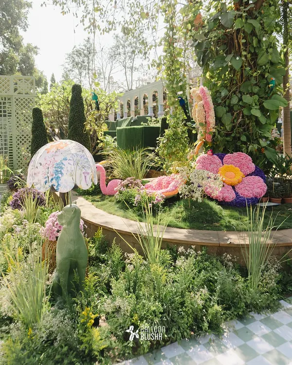 A wide view of a whimsical garden installation, featuring oversized floral sculptures, a pink flamingo, and a moss-covered dog statue.