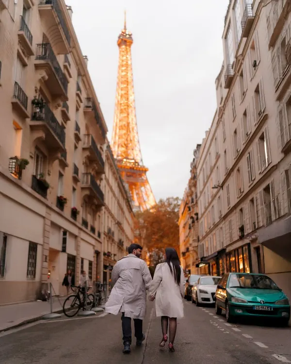 A classic Paris street scene with a romantic twist. The couple walks hand-in-hand away from the camera down a quintessential Parisian street, with the Eiffel Tower perfectly framed in the distance at golden hour.