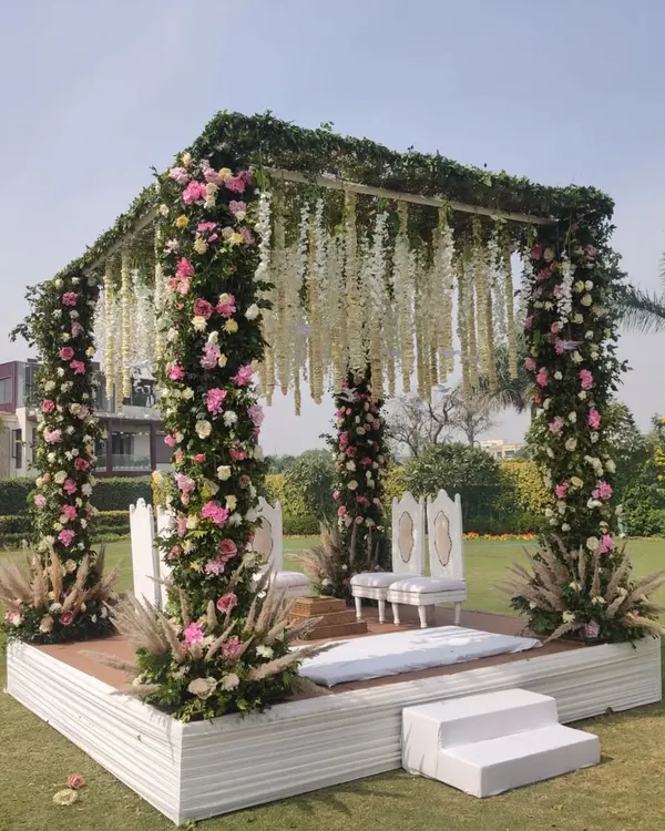 A closer look at a beautiful floral mandap for a daytime wedding. The pillars are densely wrapped with pink and white flowers, and the roof is adorned with hanging white floral strings, creating a lush, garden like feel.
