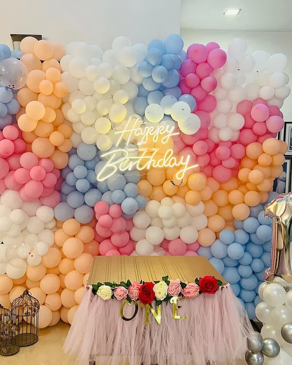 A magnificent floor-to-ceiling balloon wall in a mix of pastel colors, with a central neon 'Happy Birthday' sign. The setup is completed with a tulle-draped table for the cake.