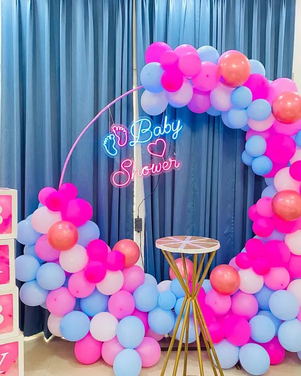 A modern baby shower setup with a circular balloon arch in pink, blue, and white. The custom "Baby Shower" neon sign with a small footprint detail adds a unique and stylish touch to the celebration.