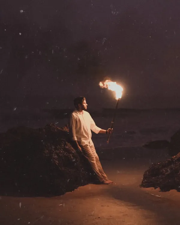A striking solo portrait of the groom-to-be, leaning against a rock on the beach at night while holding a flaming torch. The firelight creates dramatic shadows, resulting in a powerful and moody pre-wedding shot.