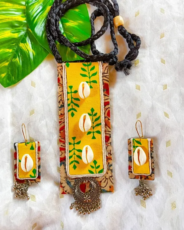 A vibrant yellow fabric panel, hand-painted with green leaf motifs and adorned with cowrie shells, is the centerpiece of this necklace. It's layered over a traditional Kalamkari print and finished with an oxidized silver charm. This is a perfect example of my layered, mixed-media style.