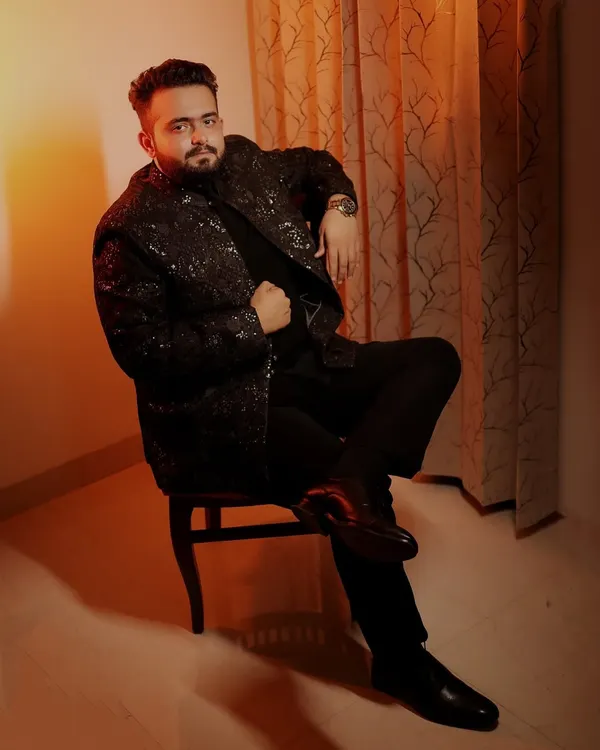 A confident, full-length portrait of the groom seated, showcasing his stylish black sherwani against a warm, lit background.