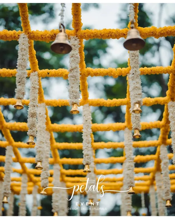 A close-up of a mandap ceiling, showcasing a beautiful grid of yellow and white chrysanthemum garlands with hanging temple bells. This detail adds sound, color, and tradition to the ceremony.
