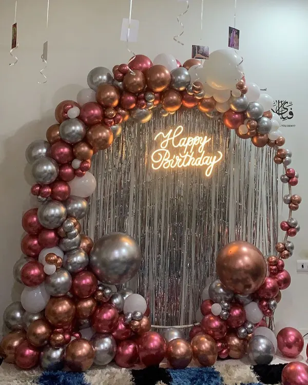 Our 'Happy Birthday' neon sign is the perfect centerpiece for this home setup, glowing against a silver fringe curtain and a metallic balloon arch.