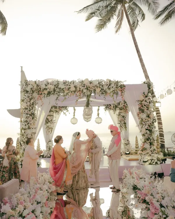 A stunning wide shot of a beach wedding ceremony at sunset. The couple stands under a magnificent floral mandap as the golden light kisses the horizon.