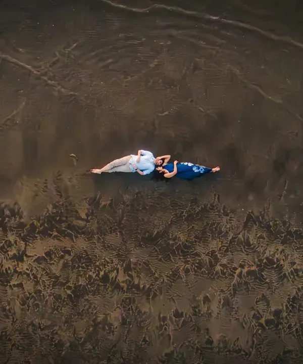 An artistic aerial shot of the couple lying on the wet sand. The patterns left by the waves create a beautiful, textured canvas, turning a simple moment into a piece of art.