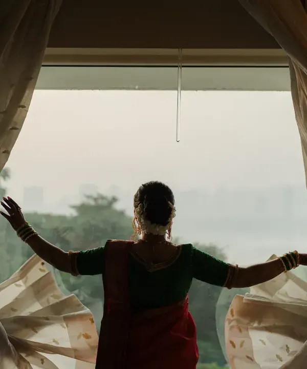 A bride looks out the window, a quiet moment of reflection before her Marathi wedding ceremony begins. The silhouette against the curtains creates a poetic and timeless bridal portrait.