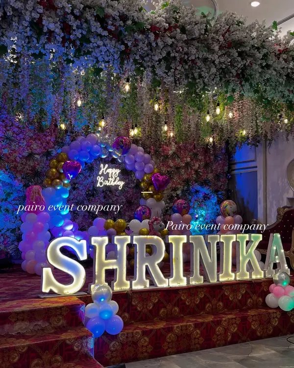A beautiful stage setup featuring large, illuminated marquee letters spelling out "SHRINIKA." The letters are the main attraction, set against a backdrop of hanging flowers and lights.
