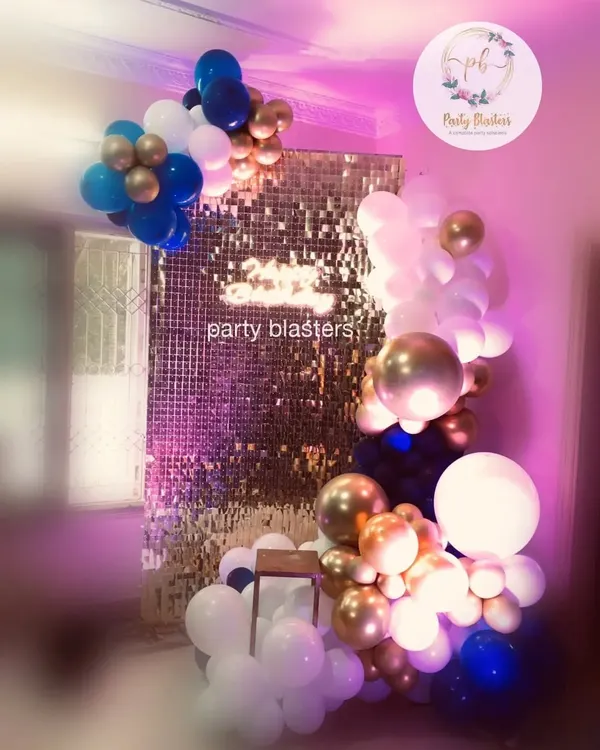 A trendy balloon garland against a gold sequin wall. We use a mix of chrome, standard, and oversized balloons in a blue, white, and gold color scheme to create a modern and glamorous photo backdrop.