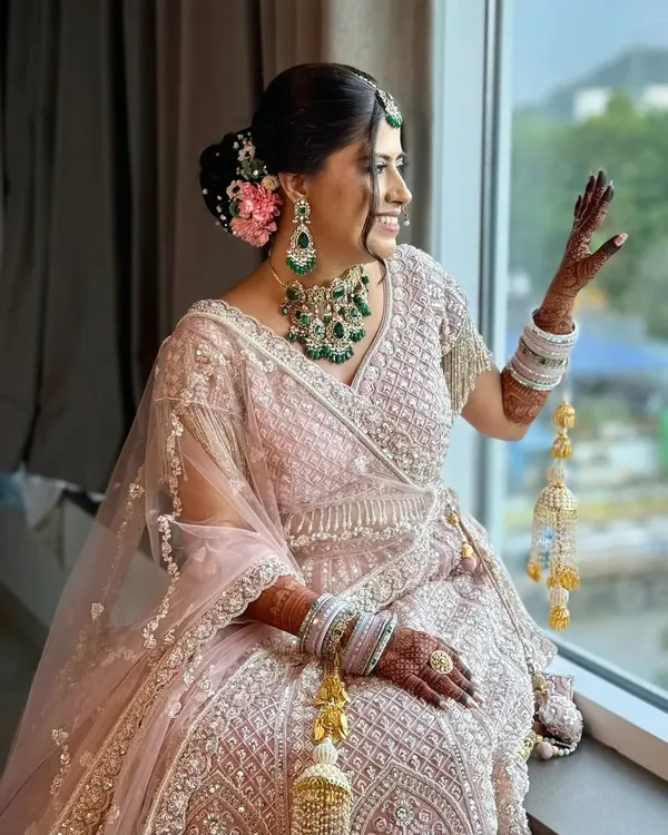 A picture of pure bridal grace. This look features soft, romantic makeup with a pop of pink in the floral bun, perfectly complementing her blush lehenga and emerald jewellery for a timeless and elegant feel.