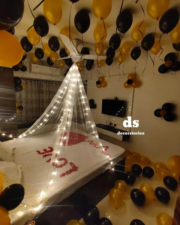 For this romantic room decoration, we installed a delicate canopy draped with fairy lights over the bed. The look was completed with black and gold balloons and a heart shape made from real rose petals.