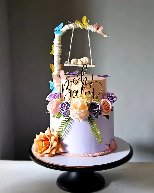 A magical baby shower cake with a swinging cradle topper. This two-tiered design in peach and lavender is adorned with sugar flowers, butterflies, and a tiny, sleeping baby handcrafted from fondant.