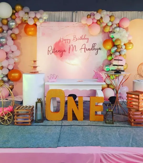 A beautiful first birthday setup for Ritanya, featuring a soft, watercolor-style backdrop. The stage is decorated with a pastel balloon garland, gold shimmer panels, and large marquee letters spelling 'ONE', creating an elegant and celebratory focal point.