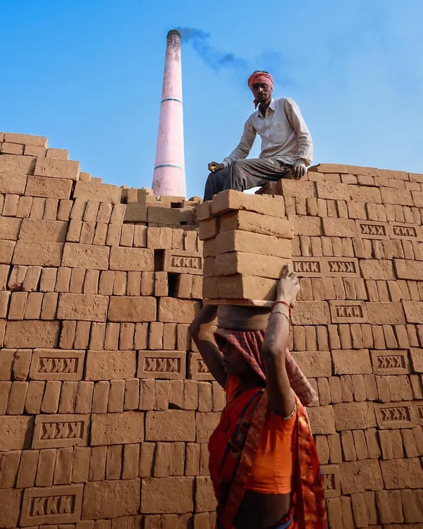 A woman carries a stack of bricks on her head while a man watches from atop a brick wall, a colorful scene depicting the daily labor at a kiln in Bihar.
