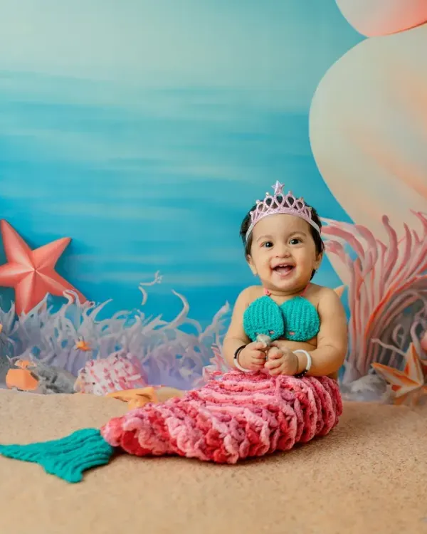 A big, happy grin from our little mermaid. The hand-knitted costume and pastel-colored 'under the sea' backdrop make this a truly magical first birthday theme.