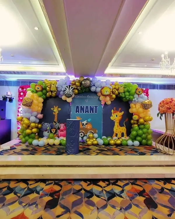 A grand jungle theme stage setup for Anant's Mundan ceremony in a banquet hall. The decor includes a large balloon arch with animal foil balloons, a personalized backdrop, and cute animal cutouts.