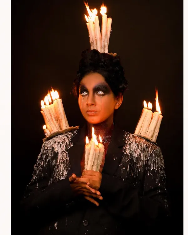 A dark fantasy portrait featuring a model with dramatic makeup, holding lit candles, and wearing a custom jacket adorned with dripping wax and more candles. This is an example of my highly conceptual and artistic work.