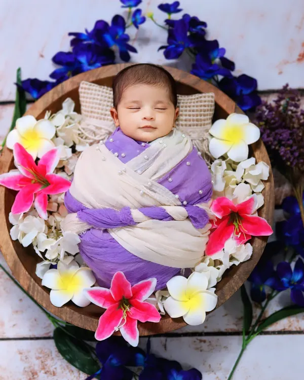 A sleeping newborn is wrapped in purple and white cloth, nestled in a wooden bowl adorned with beautiful, colorful flowers.