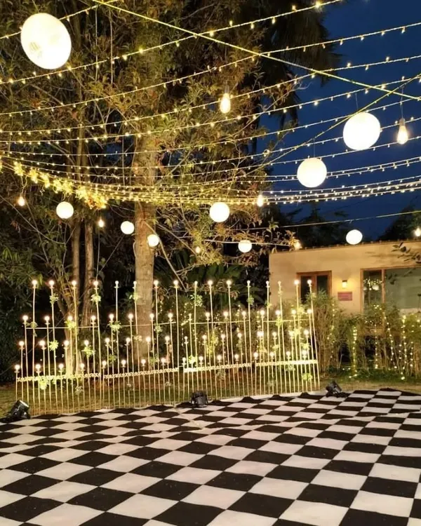 An outdoor Sangeet setup designed for a night of celebration. The checkerboard dance floor is surrounded by a fence of lights and set beneath a canopy of string lights and paper lanterns, creating a magical atmosphere.