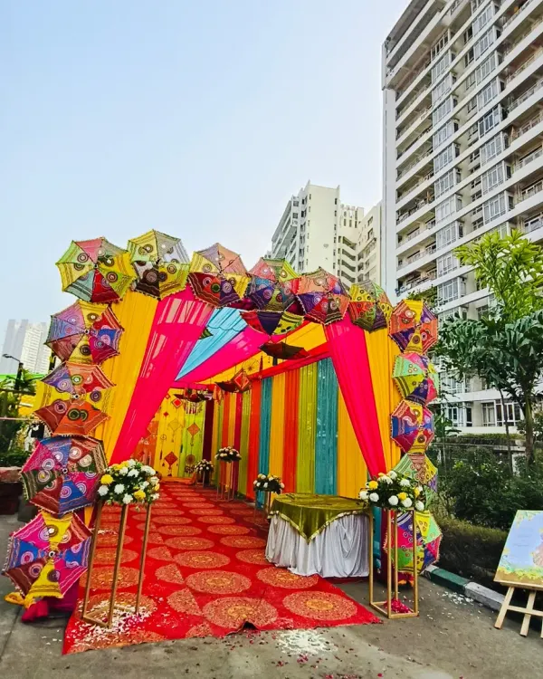 A grand and festive entrance to welcome your guests. This setup features a vibrant tunnel of pink, yellow, and blue drapes, framed by an arch made of colorful Rajasthani umbrellas, setting a joyful tone for a Mehendi or Haldi event.