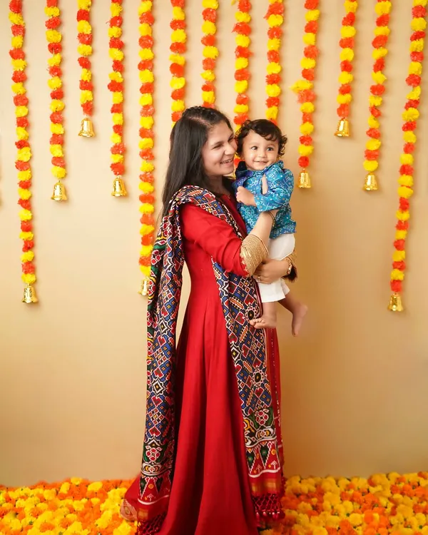 A mother in a beautiful red traditional outfit holds her son during a Ganesh Chaturthi celebration. The background is decorated with marigold garlands, creating a vibrant and festive scene.