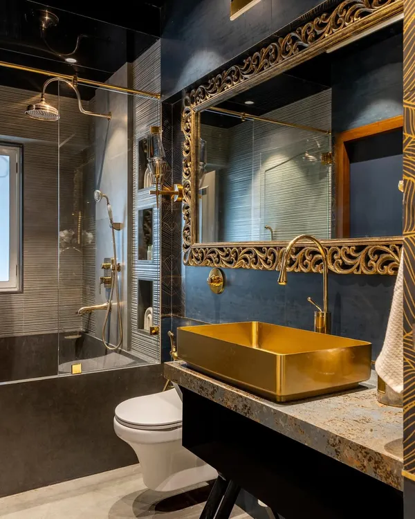 This bathroom is the epitome of opulence. A bold, gold-leafed mirror and a matching rectangular vessel sink create a dramatic focal point against the dark, textured walls and gold fixtures.