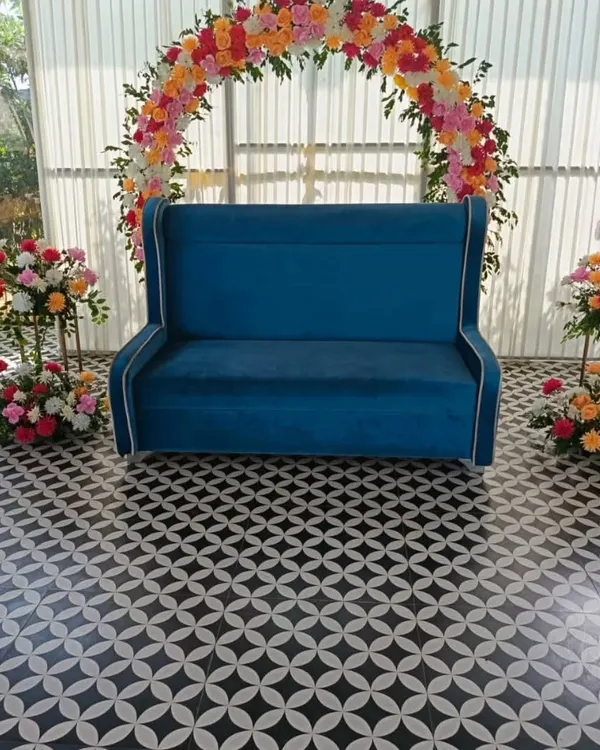 This engagement photo corner features a vibrant semi-arch of artificial flowers in shades of pink, orange, and yellow. The setup is completed with a stylish blue wingback sofa for the couple, making it a perfect spot for pictures.