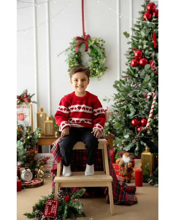 A bright smile that perfectly captures the magic of Christmas. This little boy is ready for the holidays in his festive sweater, surrounded by classic Christmas decor.