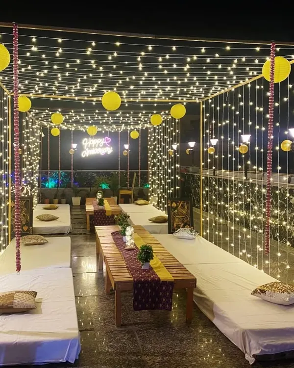 A stunning terrace anniversary setup featuring a canopy of fairy lights and yellow paper lanterns. The low wooden tables with floor seating create a cozy and intimate atmosphere for a group celebration.