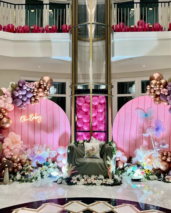 This grand baby shower setup in a spacious hall features twin pink backdrops with an 'Oh Baby' neon sign. The decoration is built around a plush guest-of-honor chair, framed by lavish balloon arrangements in shades of purple, pink, and rose gold, accented with artificial flowers and whimsical props.