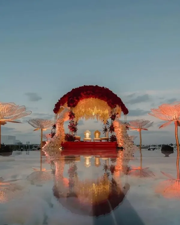 A breathtaking mandap that appears to float on water. This design features a canopy of red and white florals, with the reflection on the mirrored floor creating a truly dreamy and surreal atmosphere for a sunset ceremony.