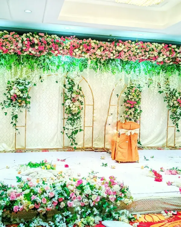 This is a classic, full-floral stage I designed for a large wedding reception. The setup includes a dense flower border, hanging green and white florals, and elegant gold-arched frames to create a grand and lush backdrop.
