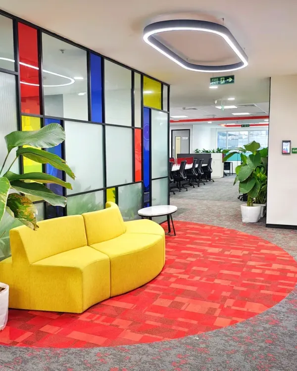 A vibrant lounge area in the Mindsprint office, featuring a curved yellow sofa set against a Mondrian-inspired colorful glass partition. The circular red carpet defines the space, making it a perfect spot for informal chats and breaks.